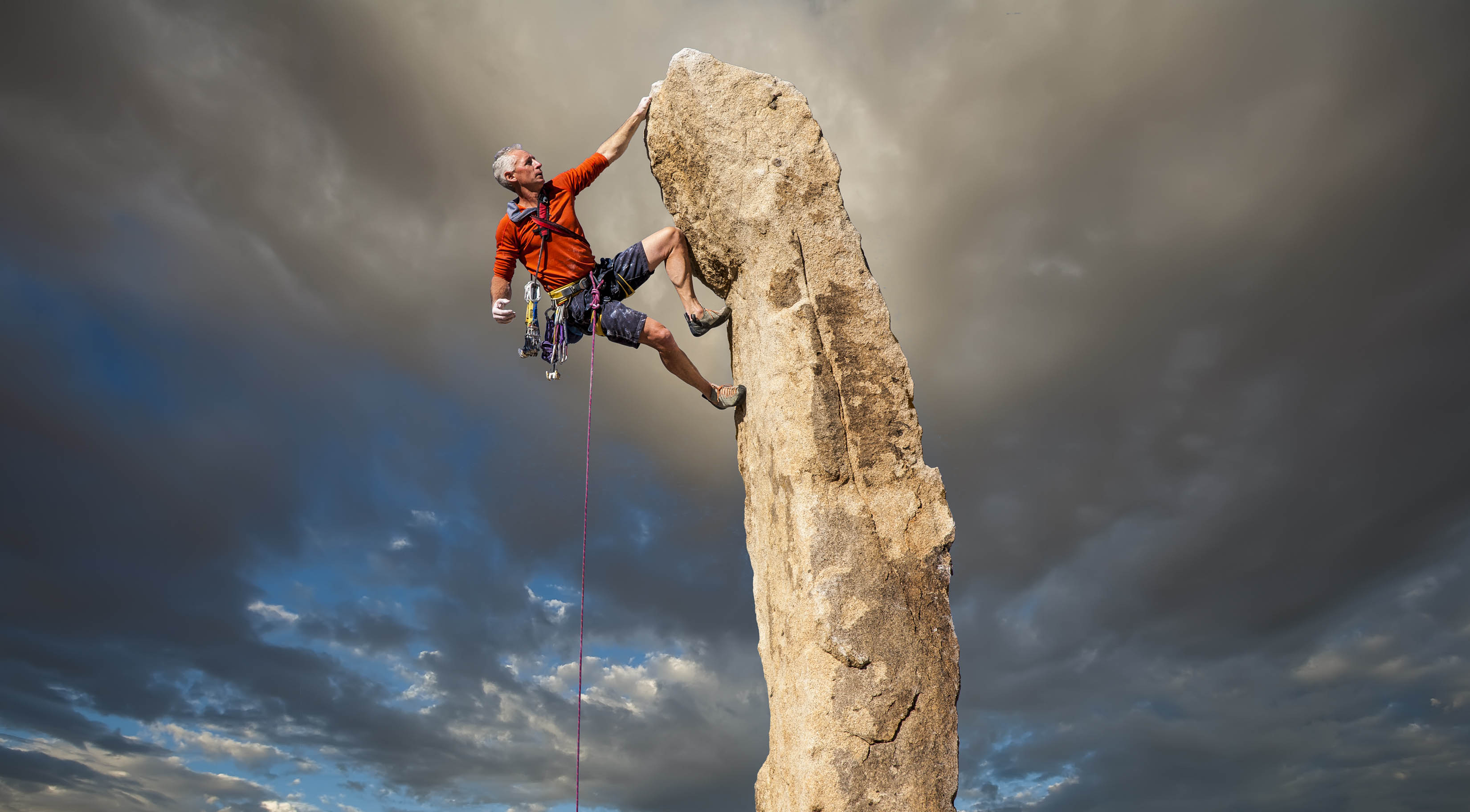 Older fit man climbing the top of a thin mountain, sky in the background is ominous